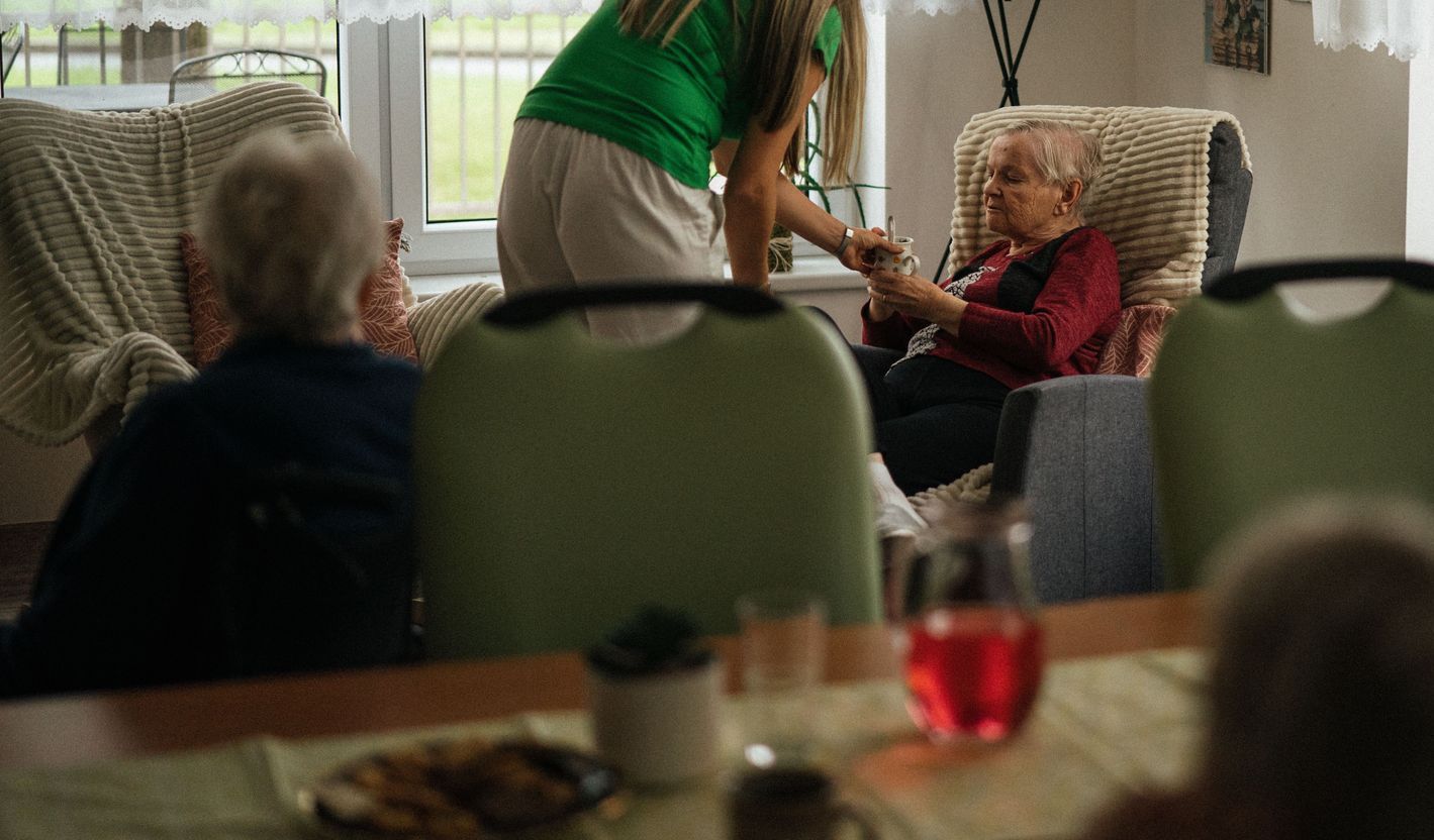 Caregiver assisting elderly women with tea in a cozy nursing home setting.
